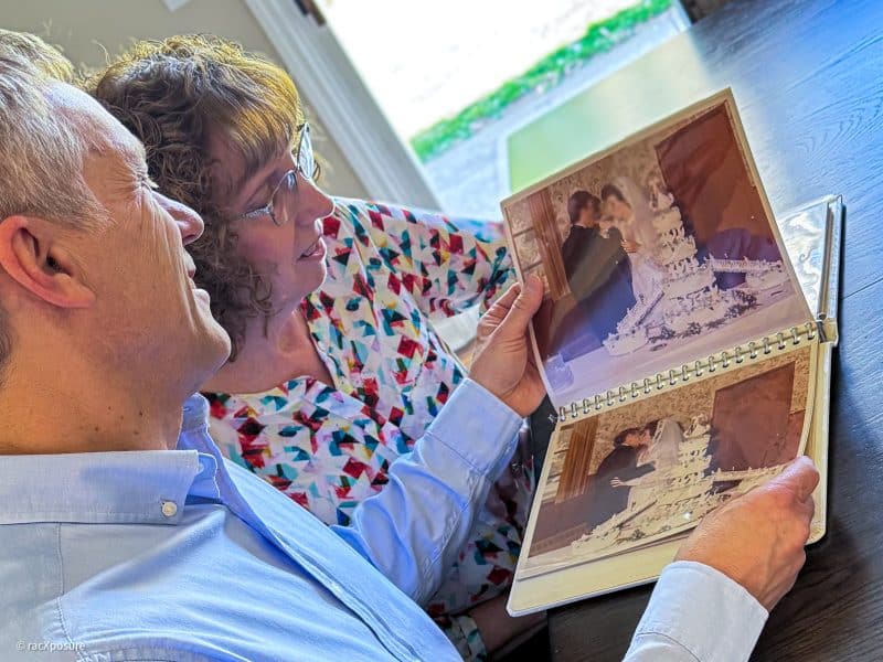A husband and wife sit down at a kitchen table and flip through their marriage album.