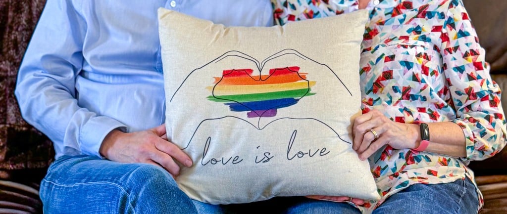 A married couple sit on a couch with a pillow between them. The pillow has an embroidered rainbow heart that says "Love is Love."