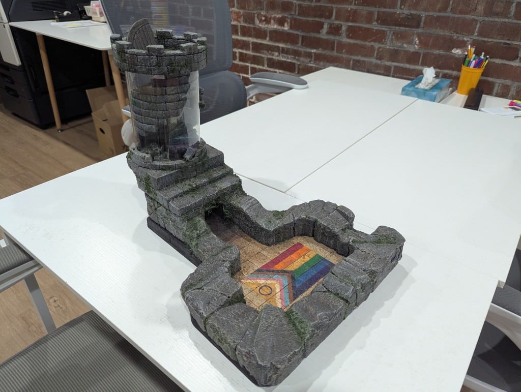 A dice tower decorated like a castle. At the end of the tower where the dice land is a Pride flag.