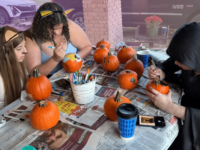 A photo of people painting pumpkins at a park's gazebo with a rainbow filter in the background.