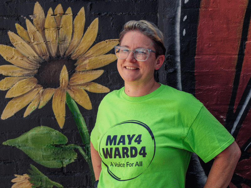 Gie May, while wearing a promotional t-shirt, poses in front of a wall with illustrations of a sun flower and butterfly.
