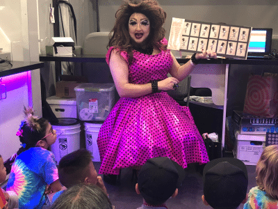 Drag queen reads a children's book to a group of children in a library.