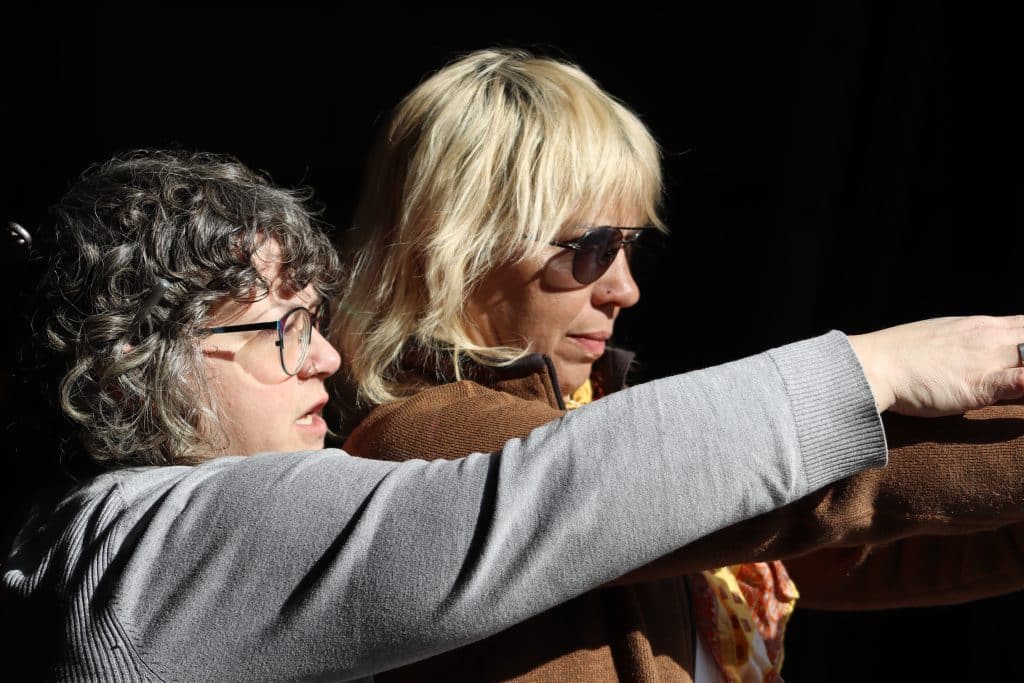 Fifi (left) raises her hand to help instruct Sarah. On the right, as sunlight streams in through the open barn door outside the frame.