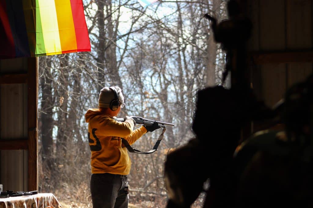The shooter in the orange jacket lowers the shotgun slightly and examines the targets from a distance. The Pride flag is barely framed, hanging beneath an open barn door.