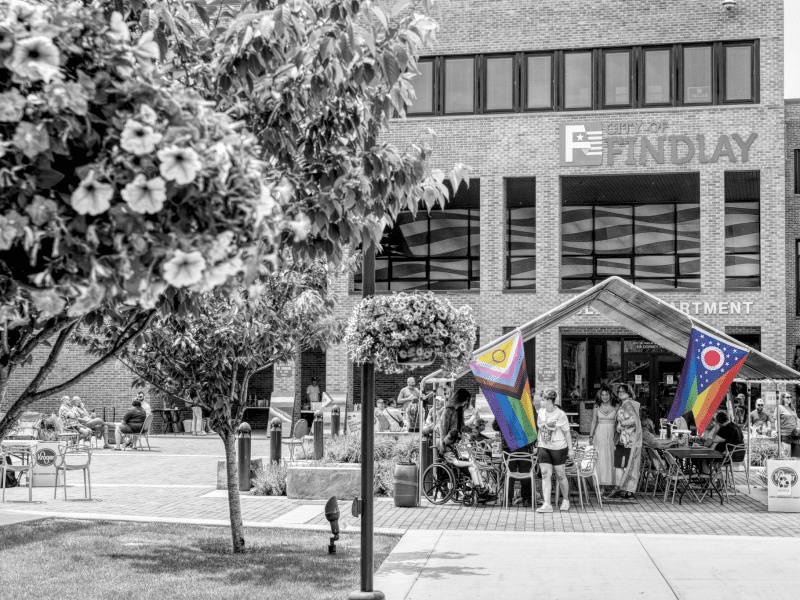 A black-and-white photo of a tent in the lower third that's in front of a Findlay, Ohio city building. Two Pride flags hang from the tent in full color, contrasting against the black-and-white.
