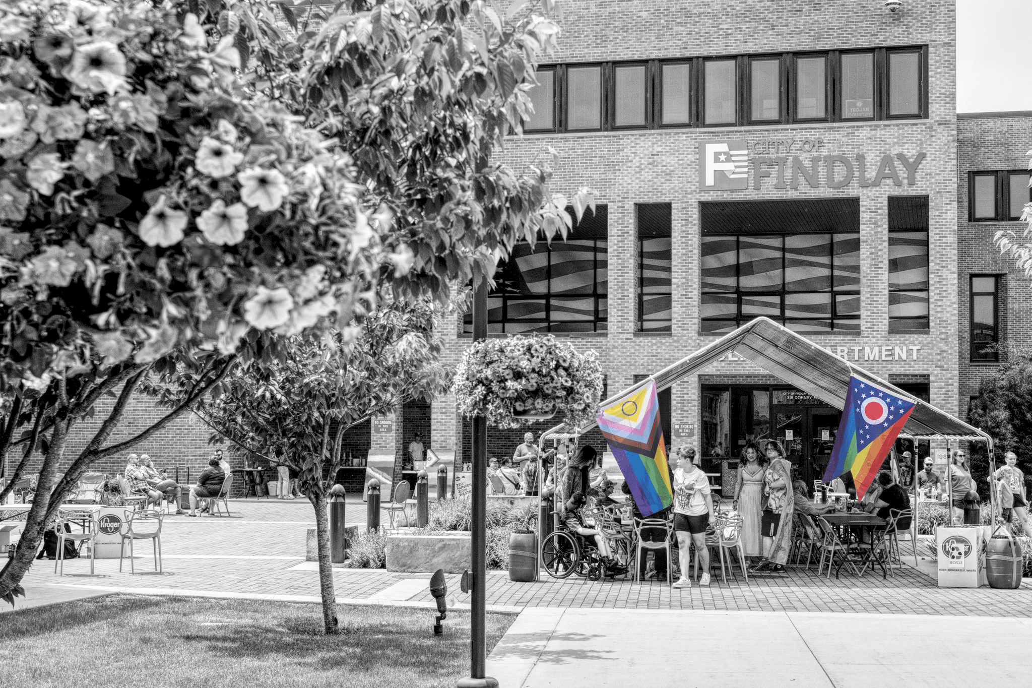 A black-and-white photo of a tent in the lower third that's in front of a Findlay, Ohio city building. Two Pride flags hang from the tent in full color, contrasting against the black-and-white.