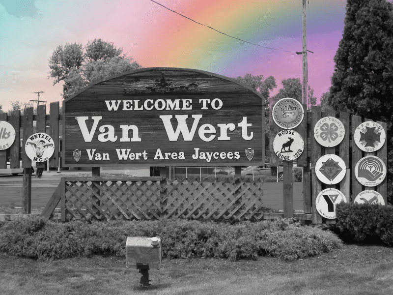 A photo of Van Wert's town sign in black and white with a rainbow superimposed in the background as if it's spilling into the image.