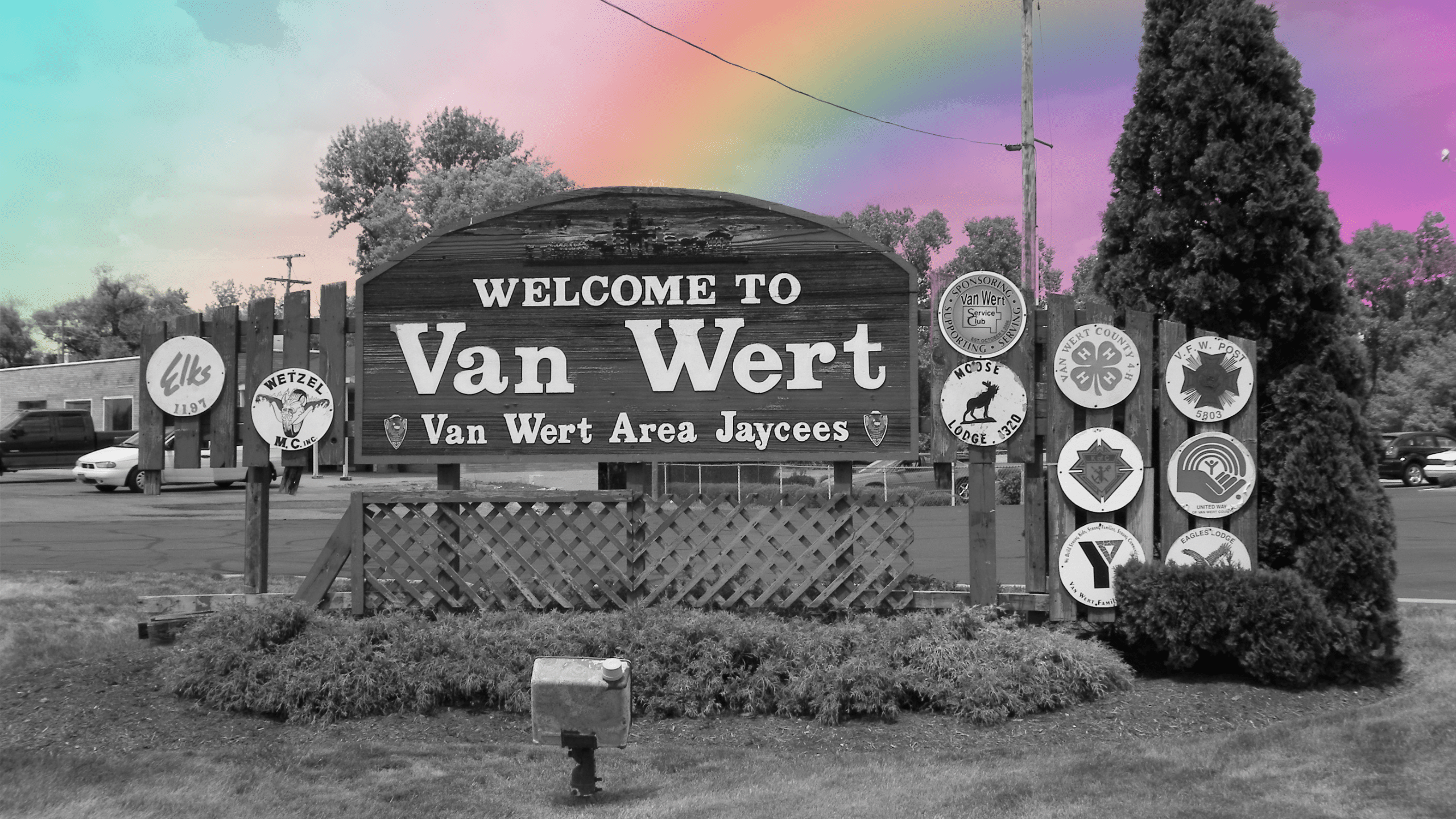 A photo of Van Wert's town sign in black and white with a rainbow superimposed in the background as if it's spilling into the image.