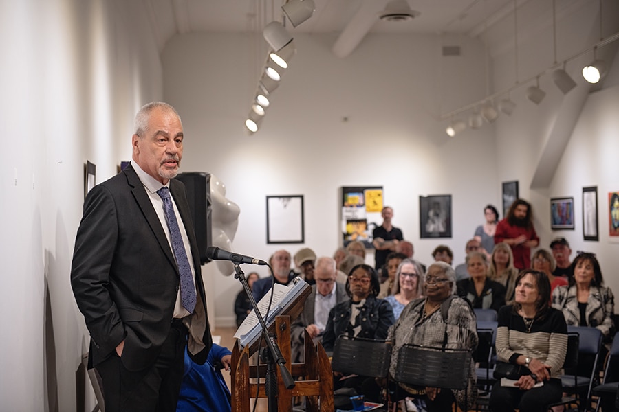 Jeff Givan, an older white man with graying hair, stands in front of a microphone and a packed room to address the audience. He has his hands in his suit pockets and his jacket is unbuttoned, revealing a light blue patterned tie.