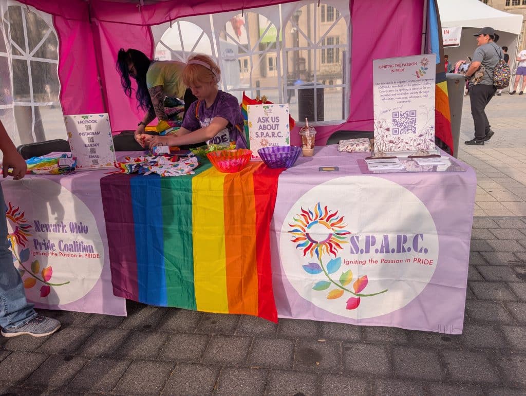 A photo of SPARC tabling at a Pride celebration. A table is draped in a pink runner showing "Newark Ohio Pride Coalition" and "SPARC." People are making crafts behind the table.
