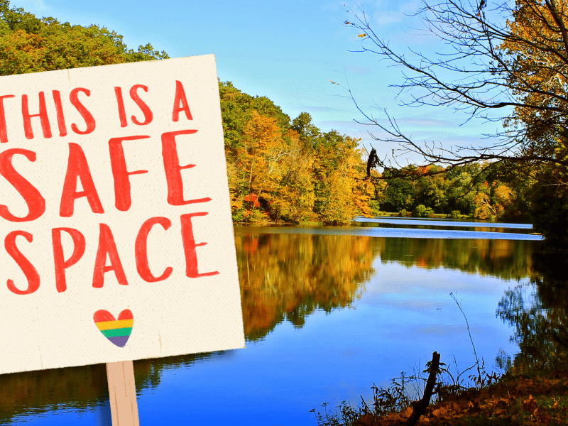 A sign is tilted as if on the bank of Glacier Lake in Youngstown, Ohio. The sign reads, "This is a safe space." A rainbow heart is underneath the lettering.