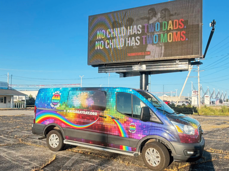 A rainbow van used by the nonprofit Have a Gay Day is parked in front of a billboard displaying an advertisement from an anti-LGBTQ+ hate group. The advertisement reads, "No child has two moms. No child has two dads."