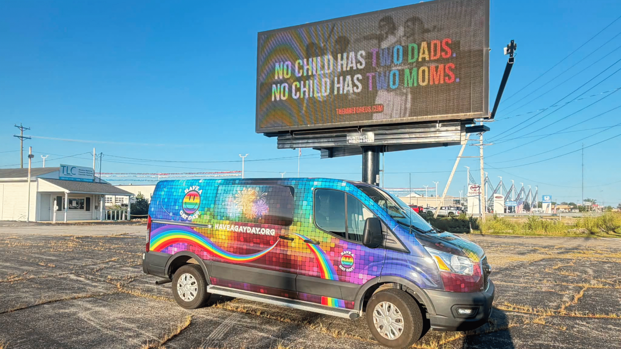 A rainbow van used by the nonprofit Have a Gay Day is parked in front of a billboard displaying an advertisement from an anti-LGBTQ+ hate group. The advertisement reads, "No child has two moms. No child has two dads."