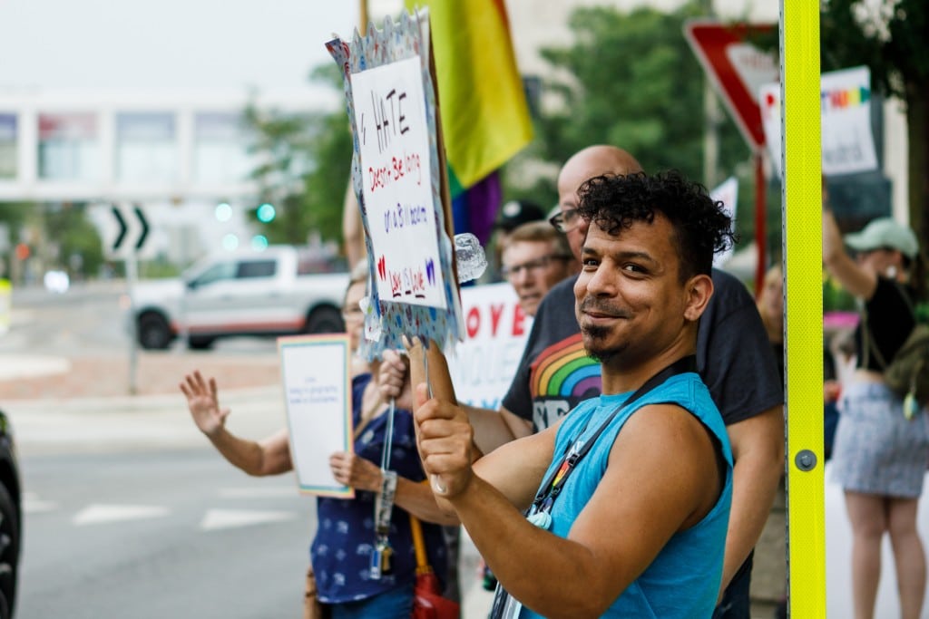 A man stands in a line of people holding a pro-LGBTQ+ sign at a rally.