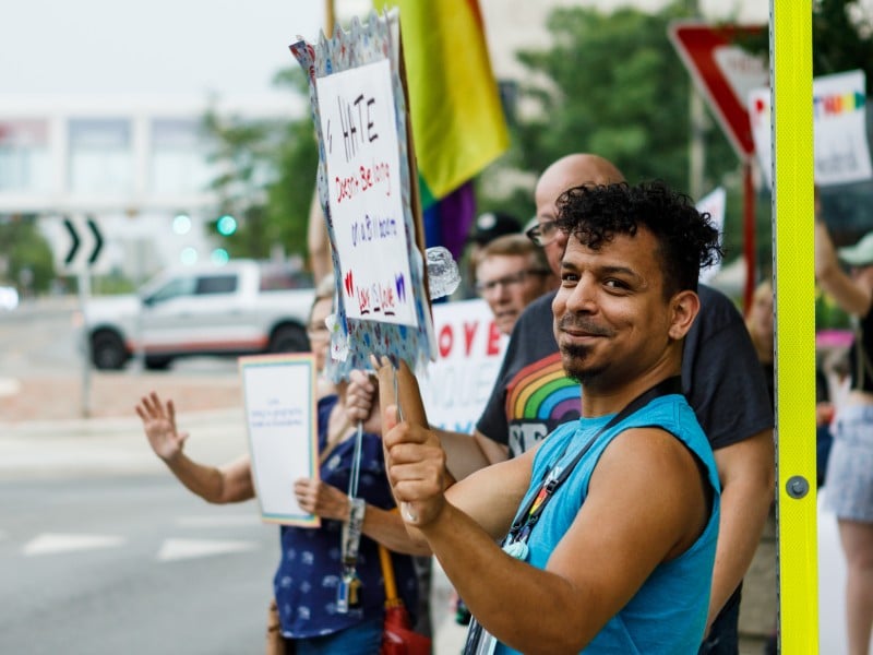A man stands in a line of people holding a pro-LGBTQ+ sign at a rally.