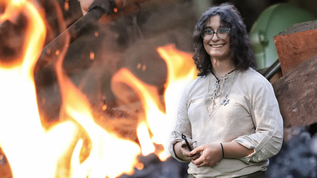 A portrait of Rose Solomon, a trans blacksmith, is superimposed in front of her sparking forge. Rose's hands are smudged with soot as she holds a leaf-like J-hook.