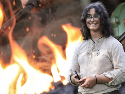 A portrait of Rose Solomon, a trans blacksmith, is superimposed in front of her sparking forge. Rose's hands are smudged with soot as she holds a leaf-like J-hook.