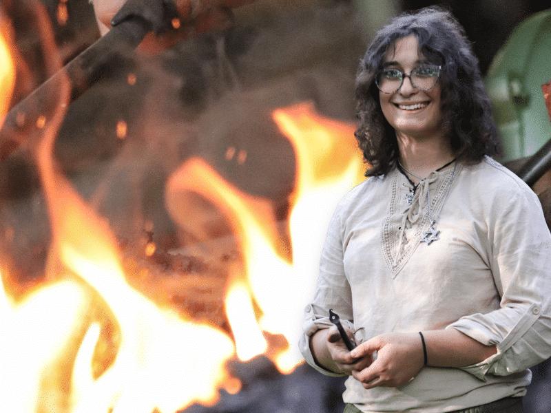 A portrait of Rose Solomon, a trans blacksmith, is superimposed in front of her sparking forge. Rose's hands are smudged with soot as she holds a leaf-like J-hook.